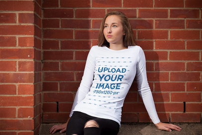Long Sleeve Tee Mockup Featuring a Serious Woman Sitting Against a Red Brick Wall 
