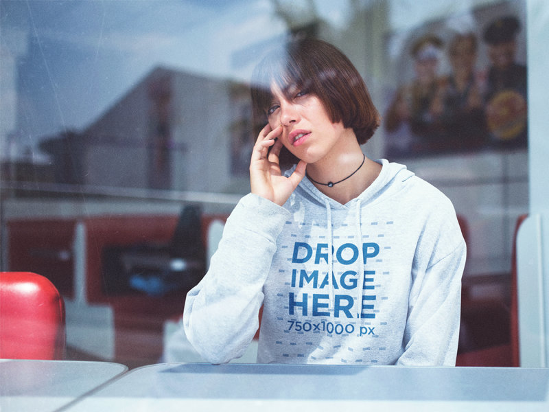 Hoodie Mockup Featuring a Young Woman Sitting Inside a Café Looking Out a Window