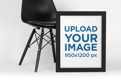 Mockup of a Framed Art Print Leaning Against a Dark Modern Chair 
