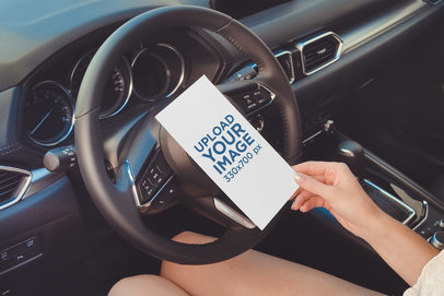 Mockup of a Woman Holding a Flyer in Front of Her Car's Steering Wheel