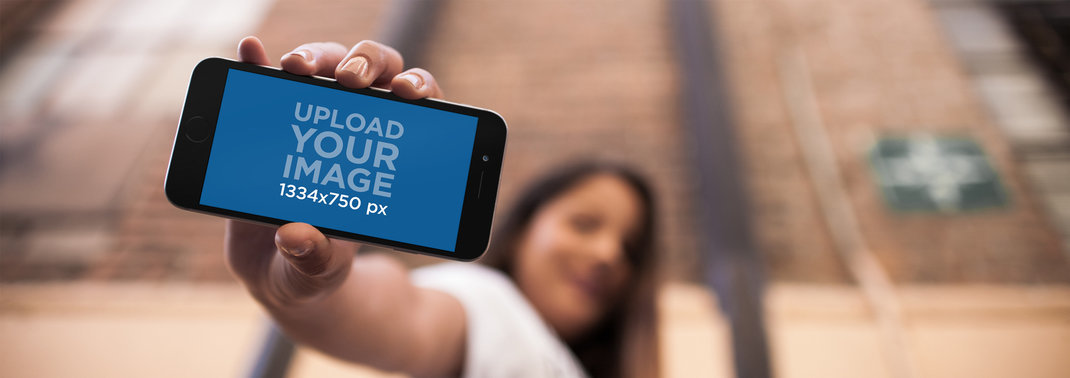 Young Beautiful Woman Holding A Black iPhone 6 In Portrait Position While At The City Mockup