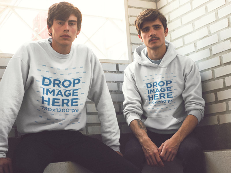 Two Serious Friends Wearing Matching Designs on a Pullover Hoodie and a Crewneck SweatshirtWhile Sitting Against a White Bricks Wall Mockup