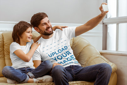 T-Shirt Mockup Featuring a Dad and His Son Sitting on a Couch and Taking a Selfie 