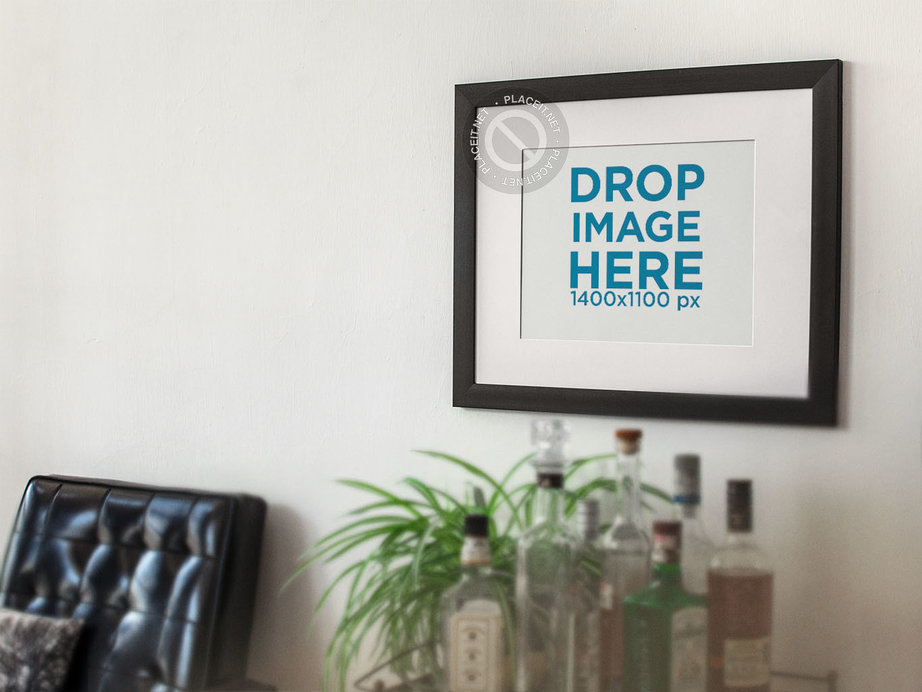 Template of a Framed Art Print on a White Wall With a Black Armchair and Bottles Nearby