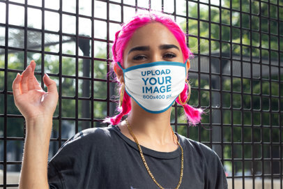 Face Mask Mockup Featuring a Young Woman With Pink Hair Posing by a Fence