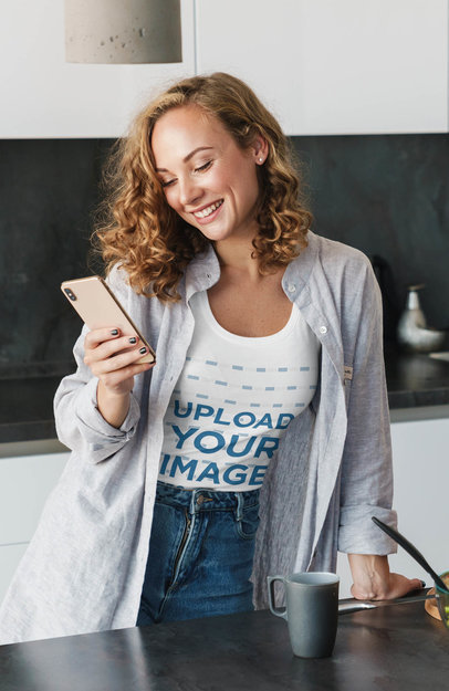 T-Shirt Mockup of a Happy Woman Using Her Smartphone in the Kitchen
