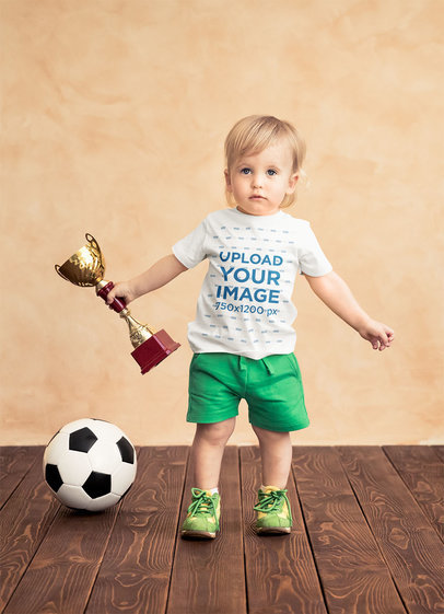 T-Shirt Mockup of a Baby Boy Holding a Soccer Trophy
