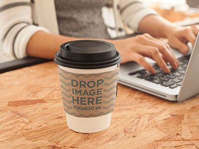 Mockup of a Coffee Cup Sitting Next to a Macbook Pro at an Office 