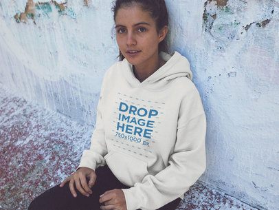 Hoodie Mockup of a Pretty Young Woman Sitting Against an Old Wall