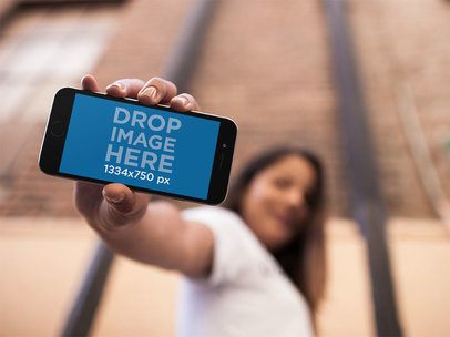 Young Beautiful Woman Holding A Black iPhone 7 In Portrait Position While At The City Mockup