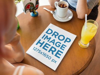 Mockup of a Magazine Lying on a Wooden Table While Two Friends are Gossiping and Having a Drink at a Coffee Shop a14365