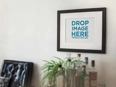 Template of a Framed Art Print on a White Wall With a Black Armchair and Bottles Nearby